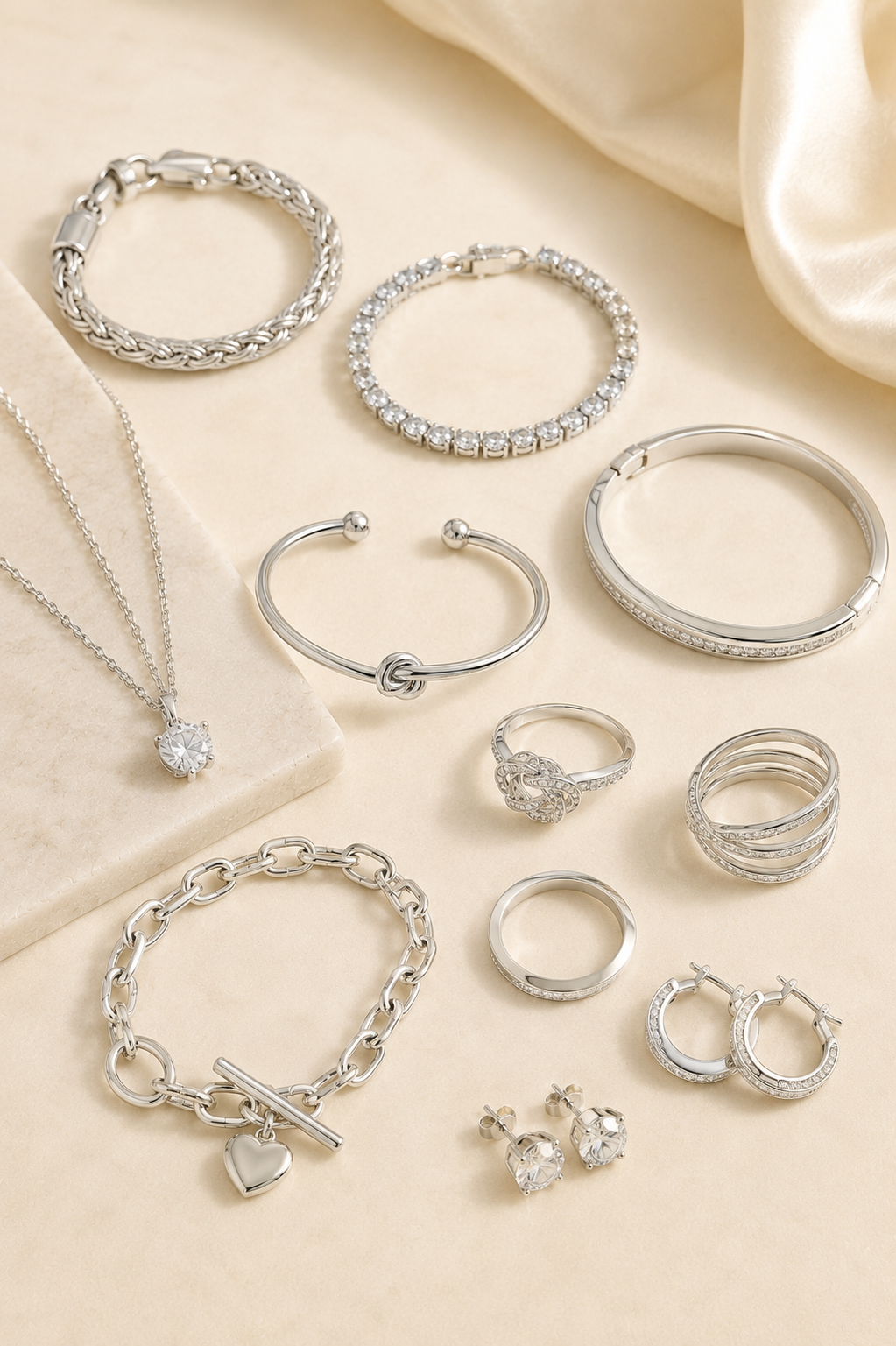 Elegant assortment of sterling silver jewelry featuring necklaces, bracelets, rings, hoop earrings, and gemstone designs displayed on a soft cream background.